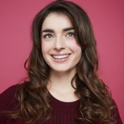 All Things Hair: Woman with romantic long brown curly hair, wearing purple jumper and posing against a pink background