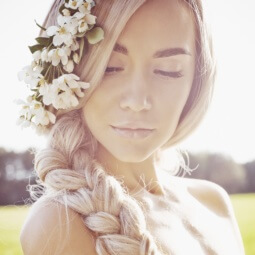 Beach wedding hair ideas: blonde model with long hair styled in a side braid with flowers placed at the top of the braid