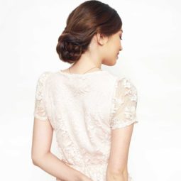 Easy prom hairstyles: Back shot of a model with chestnut brown hair styled into a low braided updo, wearing a white lace dress and posing in a studio.