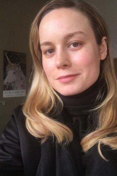 actress Brie Larson with long golden blonde hair worn in loose waves at the ends wearing an all black outfit in a room with a unicorn poster in the background