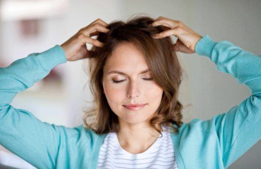 Dry scalp treatment guide: Woman with dark brown wavy medium-length hair, touching her scalp, wearing blue cardigan with white top