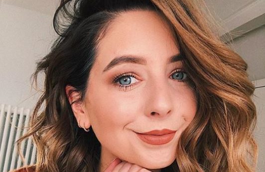 Fall hair colours: Zoe Sugg Zoella with a cinnamon wavy bob wearing a brown jumper resting her chin on her hand.