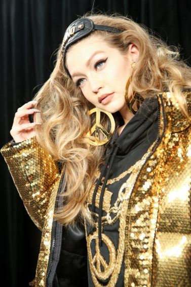 How to get thicker hair: Backstage shot of Gigi Hadid at the Moschino X H&M fashion show, with voluminous wavy blonde hair, wearing a gold puffa jacket