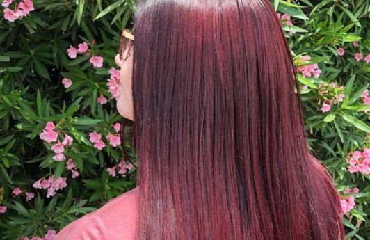 close up shot of woman with straight red violet hair, wearing pink top and posing outside next to flowers