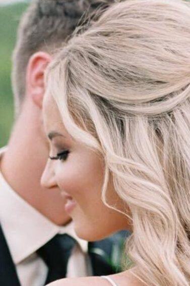 Half up half down wedding hairstyles: Woman with long bleach blonde curly styled into a half-up bun, posing outside.