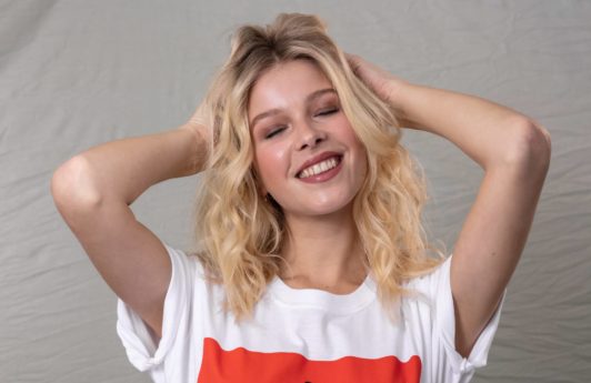 How to make hair look thicker: Blonde model with wavy shoulder length hair with her hand in her hair, wearing a white and red t-shirt