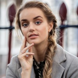 How to style long hair: Outdoor shot of a model with long blonde hair in a side fishtail braid, wearing a grey coat