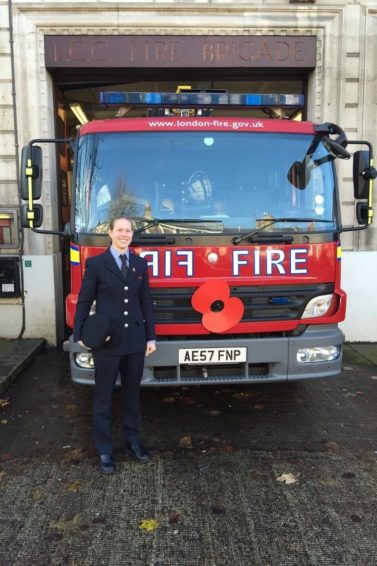 Working It: Woman with blonde hair in a bun updo standing in front of afire engine.