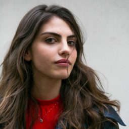 Woman with long brunette windswept hair in loose curls wearing a red top and dark jacket