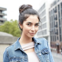 Messy bun tutorial: Brunette woman with her hair in a messy bun, wearing a denim jacket and lilac top, standing outside on the street