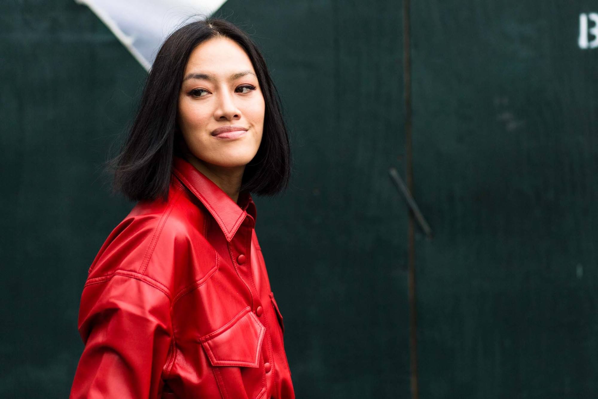 Woman with dark brown hair styled into a blunt bob, wearing red PVC outfit and posing on the street