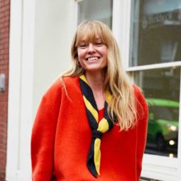 Spring haircuts: Blonde street styler with mid length wavy hair and a full fringe, wearing an orange red jumper and a striped neck scarf, standing outside in the street