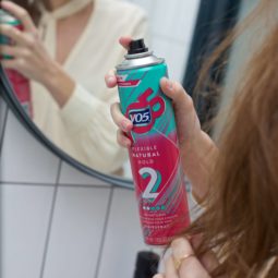 Woman spraying her brown hair with VO5 Flexible Hold Hairspray in the mirror.