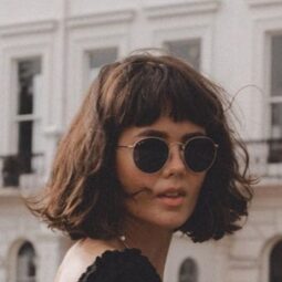 Woman with thick brown French bob wearing round sunglasses, a black top and a long denim skirt in front of a pretty white building.