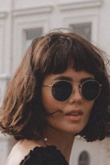 Woman with thick brown French bob wearing round sunglasses, a black top and a long denim skirt in front of a pretty white building.